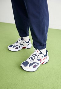 Balti ir tamsiai mėlyni sportiniai bateliai su raudonomis detalėmis, pagaminti iš akių audinio ir sintetinės medžiagos, su apvaliu pirštu, su raištelių dizainu, derinami su tamsiai mėlynomis kelnių kraštais ir baltomis kojinėmis.