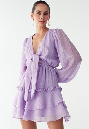 Lawendowa, lekka sukienka z głębokim dekoltem w serek, długimi bufkami na rękawach, wiązaniem w talii oraz warstwowym, falbanowym dołem. Półprzezroczysta tkanina.