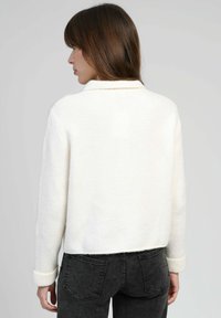 Femme aux cheveux bruns portant un pull blanc à manches longues et un jean noir, vue de dos contre un fond gris uni.