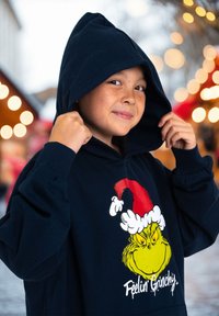 Felpa colore navy con una grande grafica cartoon del Grinch che indossa un cappello di Babbo Natale rosso. Il tessuto sembra morbido, con una tasca a marsupio e polsini a coste.