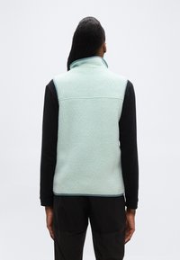 Άτομο με μακριά μαλλιά φοράει ανοιχτό μπλε υφασμάτινο γιλέκο από φλις πάνω από μαύρο πουκάμισο με μακριά μανίκια, στέκεται μπροστά σε απλό λευκό φόντο.