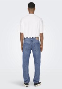 Jeans de denim azul de corte relajado con una textura desgastada, dos bolsillos traseros y una etiqueta de marca en la cintura; combinados con una camiseta blanca.