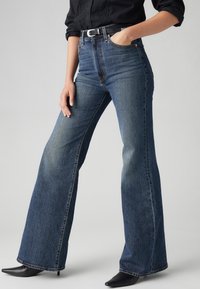 High-waisted blue denim jeans with a wide-leg cut, featuring subtle fading, contrasting stitching, and a black belt with a silver buckle.