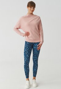 Svijetloružičasti oversized pulover s dugim rukavima uparen s plavim leggingama s uzorkom apstraktnih oblika. Bijele tenisice upotpunjuju izgled.