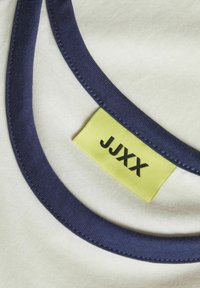 T-shirt met een crèmekleurig lichaam, een marineblauwe kraag en een gele label met "JJXX". De kraag heeft een gladde textuur en stikseldetail.