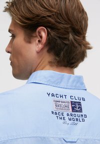 Hellblaues Hemd mit Kragen aus strukturiertem Stoff, auf dessen Rückseite ein Aufnäher mit der Aufschrift "Yacht Club" und "Race Around the World" in Dunkelblau angebracht ist.