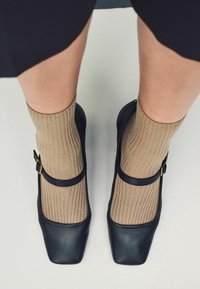 Nohy v béžových proužkovaných ponožkách s černými koženými Mary Jane botami stojí na světle šedé podlaze.