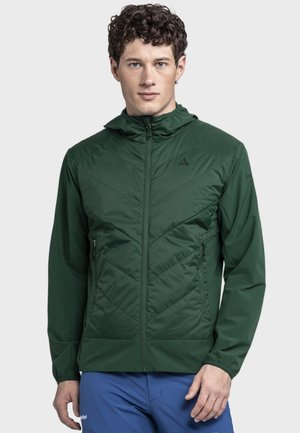 Mann trägt dunkelgrüne Jacke mit Reißverschluss und Kapuze sowie blaue Hose, steht vor schlichter hellgrauer Hintergrund.