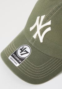 Cappellino da baseball verde oliva con logo ricamato bianco, cintura regolabile e etichetta cucita che indica "47 Clean Up". Superficie in tessuto testurizzato.