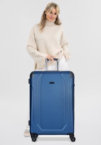 Valigia rigida blu con finitura texturizzata, manico telescopico, quattro ruote e cuciture prominenti. Design compatto per i viaggi.