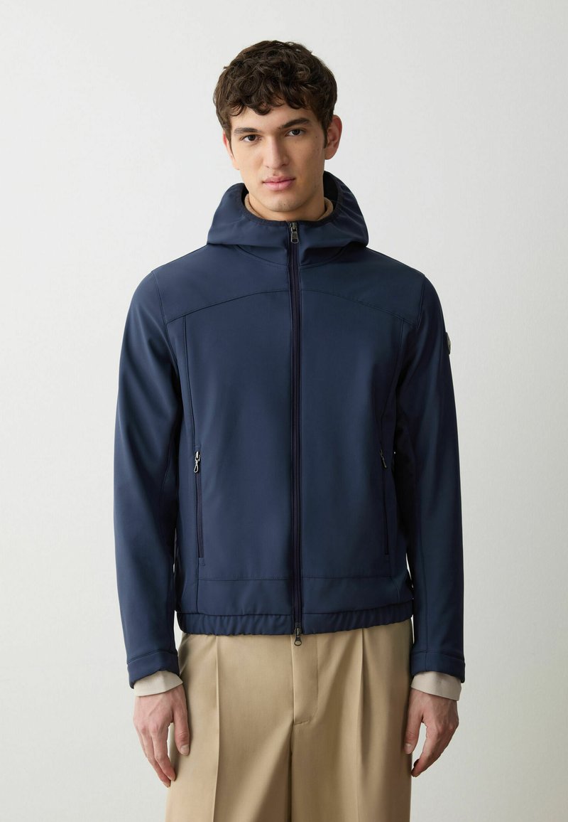 Giacca zip-up blu navy con cappuccio, realizzata in un materiale morbido. Presenta due tasche laterali con zip e una vita aderente, dal design casual.