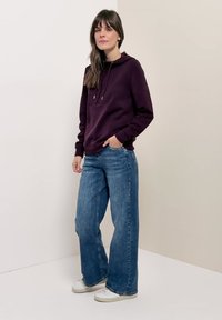 Femme portant un sweat à capuche violet foncé, un jean large bleu et des baskets blanches, debout contre un mur clair uni, une main dans la poche.