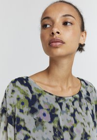 Blusa in tessuto trasparente con motivo floreale. I colori includono verde, blu e rosa tenue. Presenta un collo rotondo e maniche morbide.