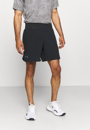 Mężczyzna w szarej koszulce z wzorem moro, czarne krótkie spodenki sportowe z logo na lewej nodze oraz białe buty Nike, stojący na białym tle.