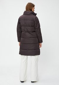 Mujer con un abrigo acolchado largo de color marrón oscuro y cuello alto, pantalones anchos blancos, de pie de espaldas sobre un fondo claro y sencillo.