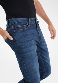 Tummansiniset farkut, joissa on slim fit -malli, napin ja vetoketjun sulkeminen, vyölenkit sekä kontrastivärinen tikkauksen yksityiskohta saumoissa.