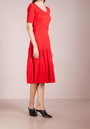 Vrouw die een rood, knielang jurkje met korte mouwen en een gelaagde rok draagt, en zwarte muilen met een hoge hak en puntige neus, staand op een lichte vloer.