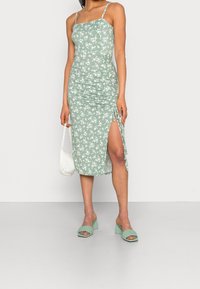 Groene bloemenjurk met een aansluitend ontwerp, geraffineerde zijkanten en een hoge split tot boven de dij. Gecombineerd met gevlochten groene sandalen met hakken en een witte tas.