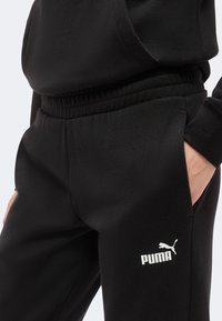 Άτομο που φοράει μαύρη φόρμα Puma με ελαστική μέση και το χέρι στην δεξιά τσέπη, δείχνοντας το λογότυπο Puma στον αριστερό μηρό.