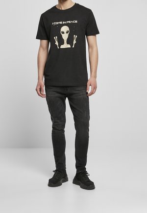 Black cotton t-shirt featuring an extraterrestrial graphic with the text "I COME IN PEACE." Paired with black slim-fit jeans and black sneakers.