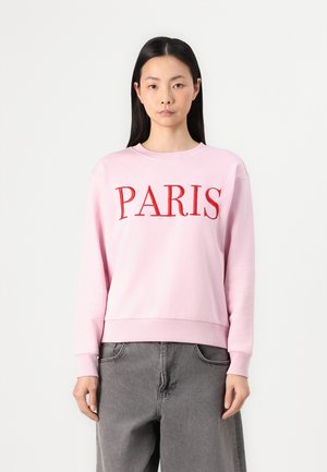 Pink sweatshirt with red "PARIS" text, round neckline, ribbed cuffs, and hem. Worn with gray baggy pants. Simple, soft cotton material.