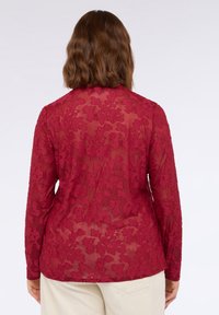 Transparente bordeauxfarbene Bluse mit floralem Spitzenmuster, langen Ärmeln und abgerundetem Saum. Das Material wirkt zart und leicht.