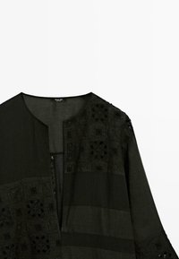 Černá halenka s dlouhým rukávem s patchworkovým designem obsahujícím vyšívané čtvercové vzory a kulatým výstřihem bez viditelných knoflíků.