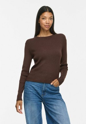 Femme aux longs cheveux foncés portant un pull côtelé marron et un jean bleu, debout avec une main dans la poche sur fond blanc.