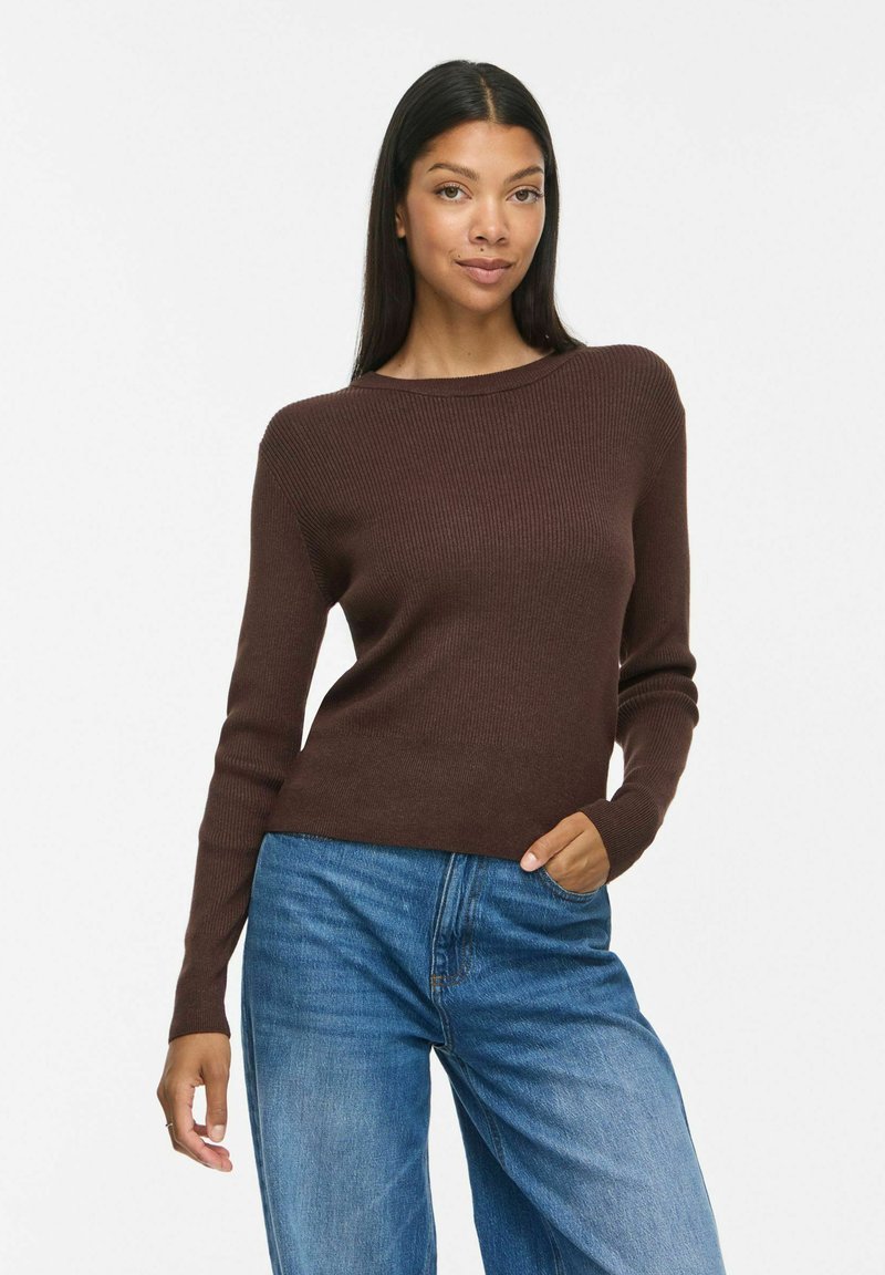 Femme aux longs cheveux foncés portant un pull côtelé marron et un jean bleu, debout avec une main dans la poche sur fond blanc.