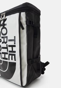 Czarny i biały plecak z trwałego materiału, z dużym logo, górnym zamknięciem na zamek, bocznymi klamrami i regulowanymi paskami. Teksturowana powierzchnia.