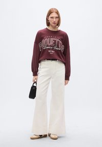 Μαύρη μπλούζα με την επιγραφή "PROUETTES Dance Team", κρεμ παντελόνι με φαρδιά μπατζάκια, μπεζ παπούτσια και μικρή μαύρη τσάντα χειρός. Λείο ύφασμα, καθημερινό σχέδιο.