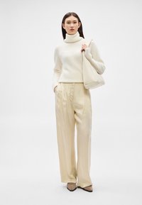 Γυναίκα που φοράει εκρού πουλόβερ με ζιβάγκο, μπεζ παντελόνι με φαρδιά μπατζάκια, παπούτσια ταουπέ με μυτερή μύτη, κρατώντας μεγάλη εκρού τσάντα ώμου, σε λευκό φόντο.