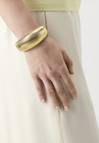 Rante dengan gelang emas lebar yang bercorak terletak di atas kain berlipat krim dan atasan kuning muda.
