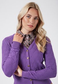 Lila stickad cardigan med ett diamantsmönster, ribbade ärmslut och knappar i guld färg. Kombination med en mönstrad skjorta med krage under.