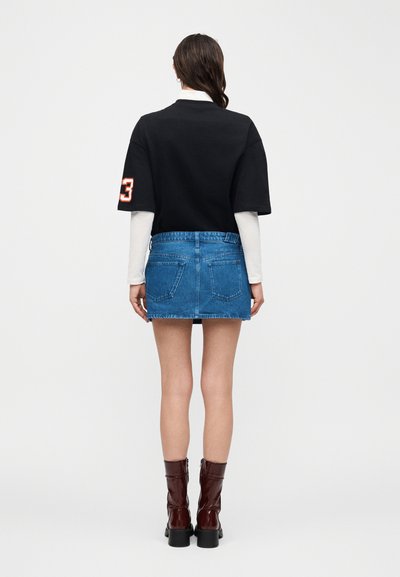 Kvinne sett bakfra iført svart kortermet topp over hvite langermede trøyer, blå denim miniskjørt og brune støvler til midt på leggen mot hvit bakgrunn.