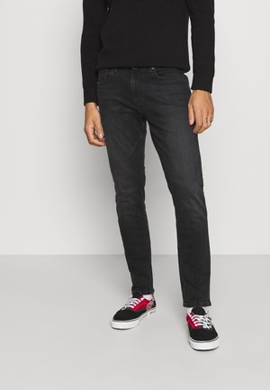 Man draagt zwarte jeans, zwarte trui en zwart-rode sneakers met witte veter, staand tegen een effen lichte achtergrond.