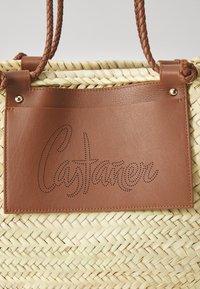 Υφασμένη τσάντα από ψάθα με καφέ δερμάτινη επιφάνεια που διαθέτει περφορημένο κείμενο. Οι πλεκοί δερμάτινοι ιμάντες συμπληρώνουν την φυσική υφή της ψάθας.