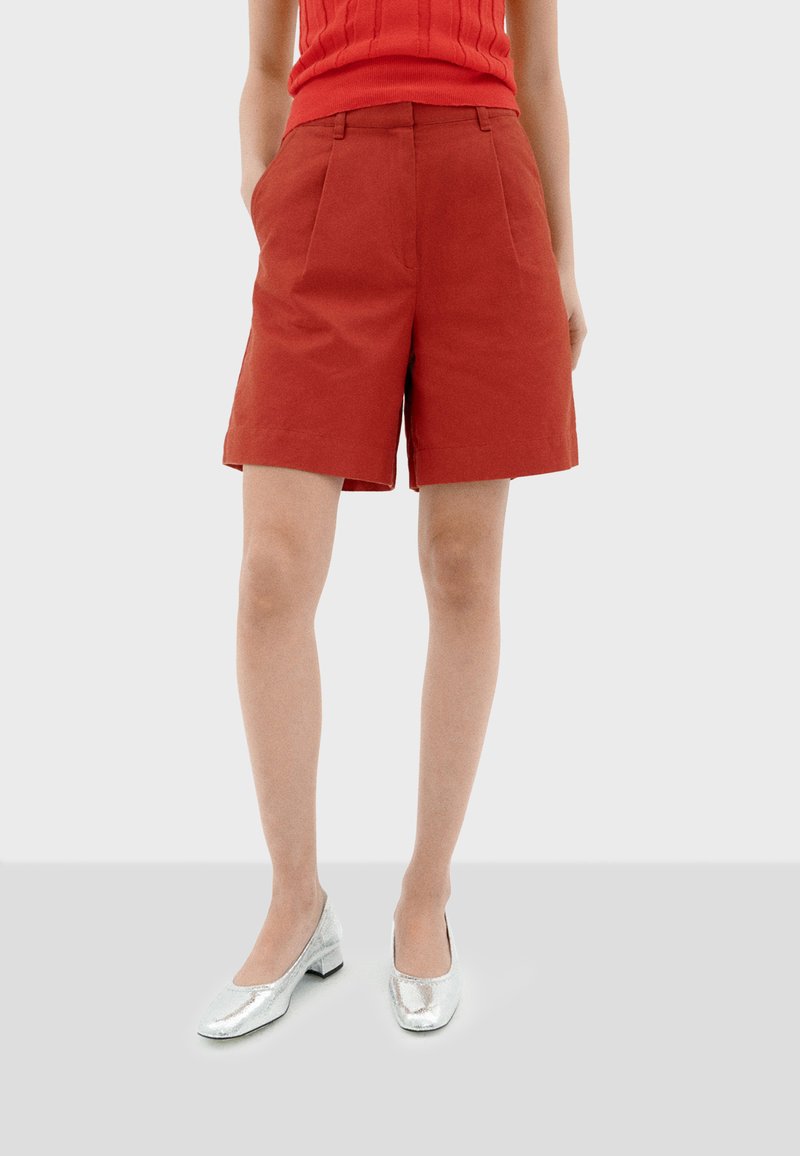 Shorts en coton rouge avec une coupe décontractée, présentant des plis et des poches latérales, associés à des ballerines en argent brillant.
