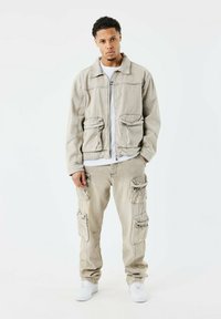 Lys beige denimjakke med to forlommer og snorede detaljer, parret med matchende cargo-bukser med flere sidelommer. Hvide sneakers.