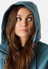 Giacca impermeabile di colore teal con cappuccio foderato e zip nera. Il materiale appare liscio con una leggera brillantezza. Tra le caratteristiche c'è un cordino al collo.