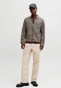 Graue, gemusterte Jacke mit durchgehendem Reißverschluss und Brusttasche, über einem burgunderroten Hemd getragen. Beige, gerade geschnittene Hose und schwarze Schuhe komplettieren das Outfit.