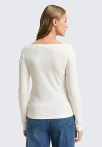 Langarmtop van zacht, ribbelwit materiaal. Heeft een vierkante halslijn en een slim fit, gecombineerd met blauwe jeans. Details omvatten minimale naden.