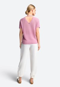 Rosa gestrickter Kurzarmpullover mit einem V-förmigen Rückenausschnitt, kombiniert mit lockeren weißen Hosen und braunen Plateausandalen.