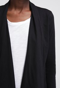 Svart cardigan med öppen frontdesign, tillverkad av mjukt tyg, lager på lager över en vit t-shirt. Har långa ärmar och en slät textur.