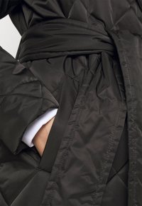 Ruka se vklouzává do kapsy černého prošívaného zimního kabátu s páskem, viditelné textury kabátu a detaily švů.