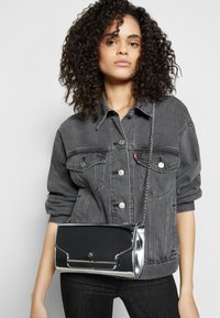 Svart lackskinnsväska med silverdetaljer, rektangulär form och kedjeband, bärs över en grå denimjacka.