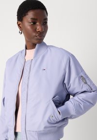 Svijetloplavi bomber jakna s rebrastim ovratnikom, džepovima na patentni zatvarač i malim brendiranim logotipom s lijeve strane. Glatka, podstavljena tekstura.