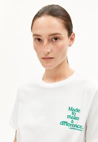 Femme avec les cheveux tirés en arrière portant un t-shirt blanc avec le texte vert "Fait pour faire une différence - Armedangels" sur un fond blanc simple.