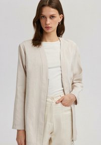 Beige letvægtsjakke med en afslappet pasform, lange ærmer og lynlås foran. Bæres over en hvid top og parret med lyse bukser.