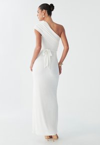 Kobieta w dopasowanej białej sukience z jednym odsłoniętym ramieniem i przewiązanym w pasie stoi na jasnym tle, prezentując eleganckie kolczyki.